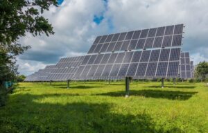 Rows of solar panels installed in a grassy field under a partly cloudy sky, surrounded by trees.